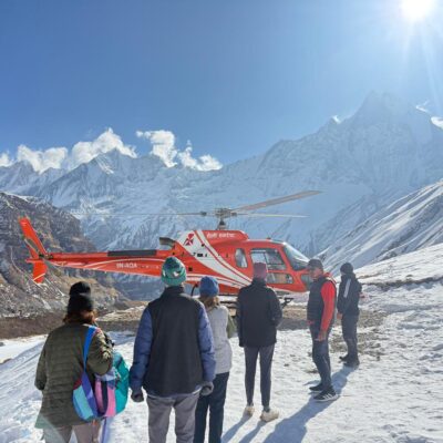 A bright red helicopter landed on a snowy mountain plateau in the Annapurna region, with a group of bundled-up travelers standing nearby and dramatic snow-covered Himalayan peaks rising under a clear blue sky.